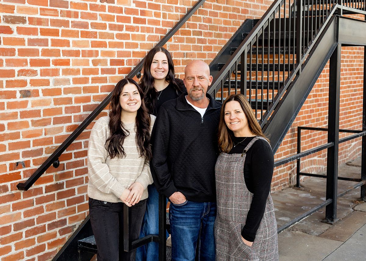 A family of four smiling together outdoors against a brick wall near a metal staircase.