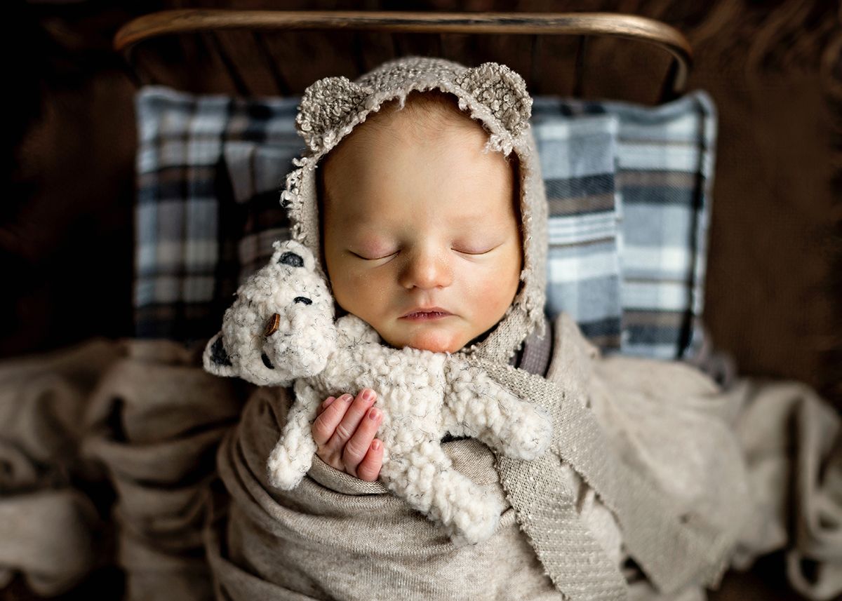Sleeping newborn wrapped in gray swaddle wearing bear ear bonnet, cuddling a soft teddy bear toy.