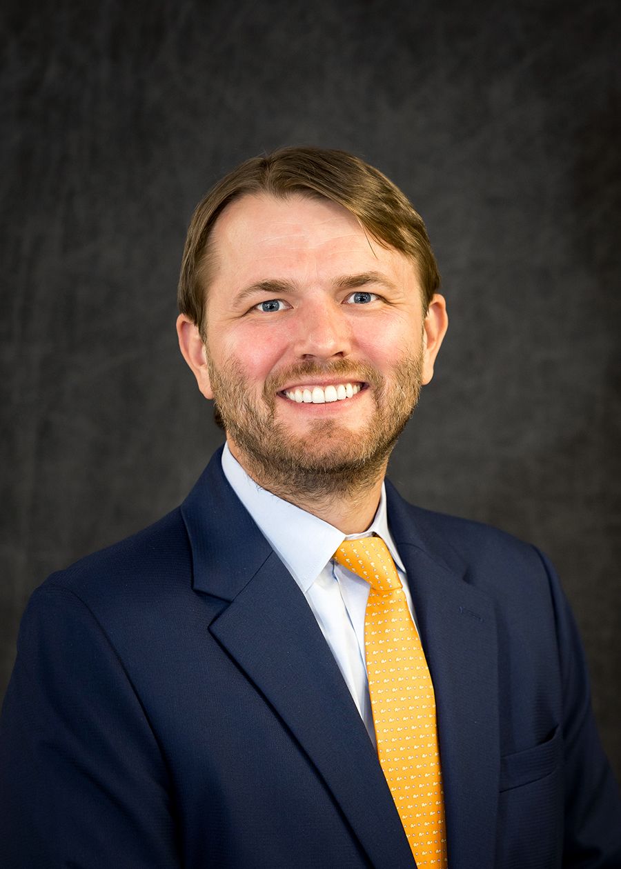 Young professional man with a beard wearing a navy blazer and gold tie smiling against a gray background.