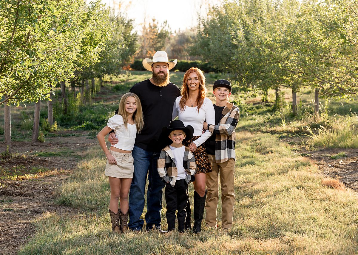 Family of five posing in a green orchard pathway at sunset.