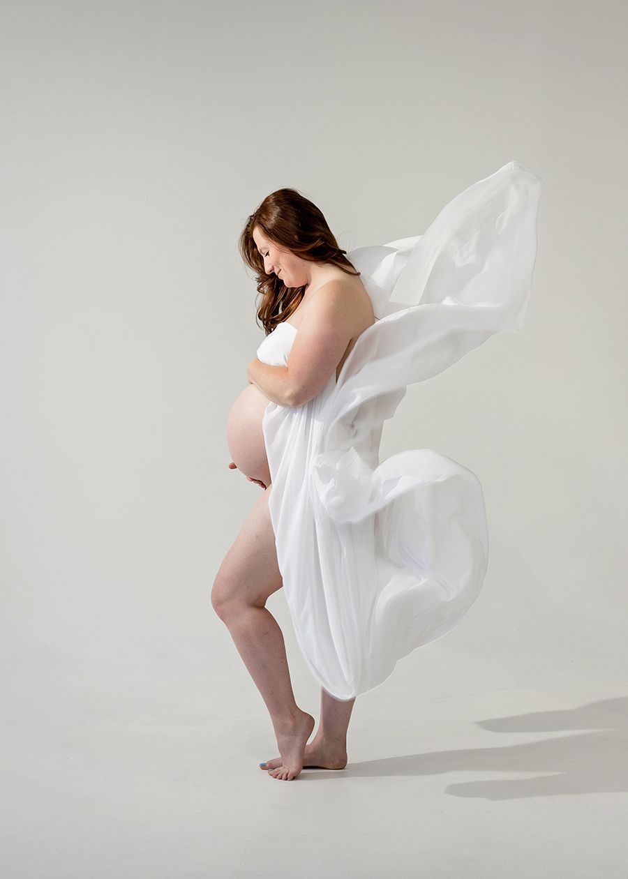 Pregnant woman draped in flowing white fabric poses gracefully in a studio maternity photography session.