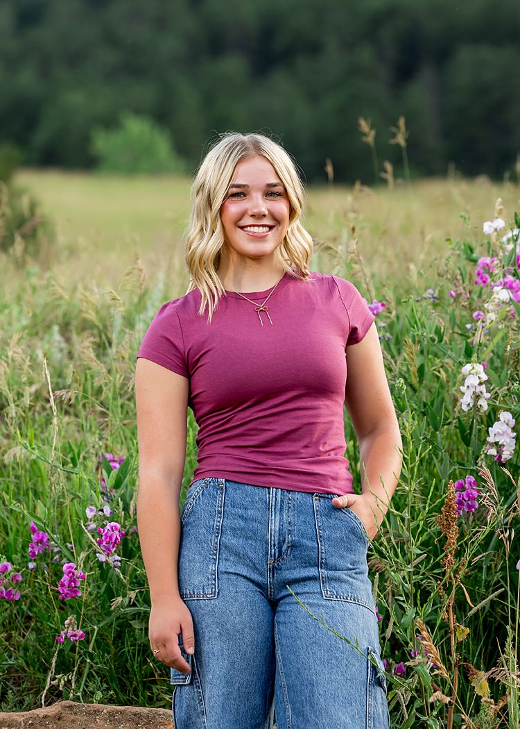Smiling blonde woman in maroon t-shirt and jeans standing in a wildflower meadow.