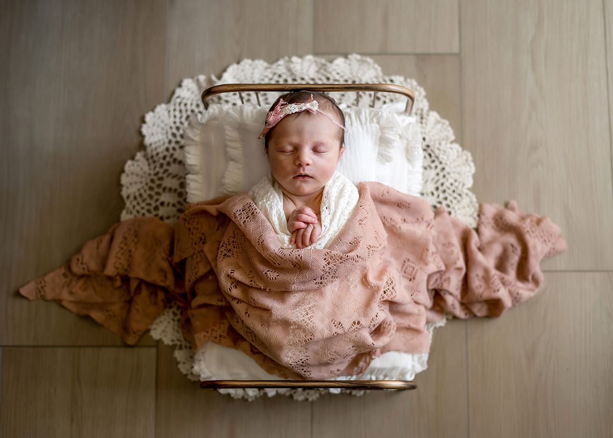 Newborn baby girl sleeping peacefully wrapped in pink lace swaddle on white doily in small wooden basket.