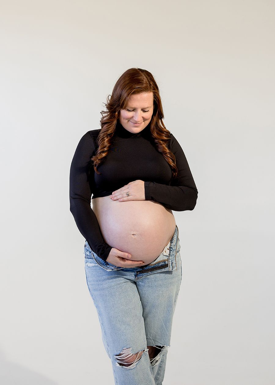 Smiling pregnant woman in black turtleneck and ripped jeans cradling her bare baby bump against a white background.