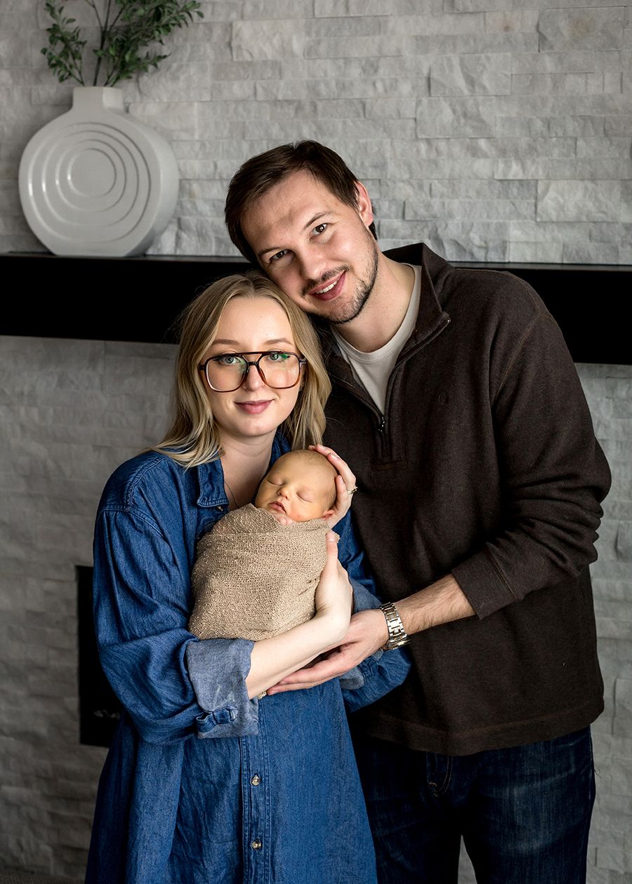 New parents smiling and holding their sleeping newborn baby in front of a stone fireplace.
