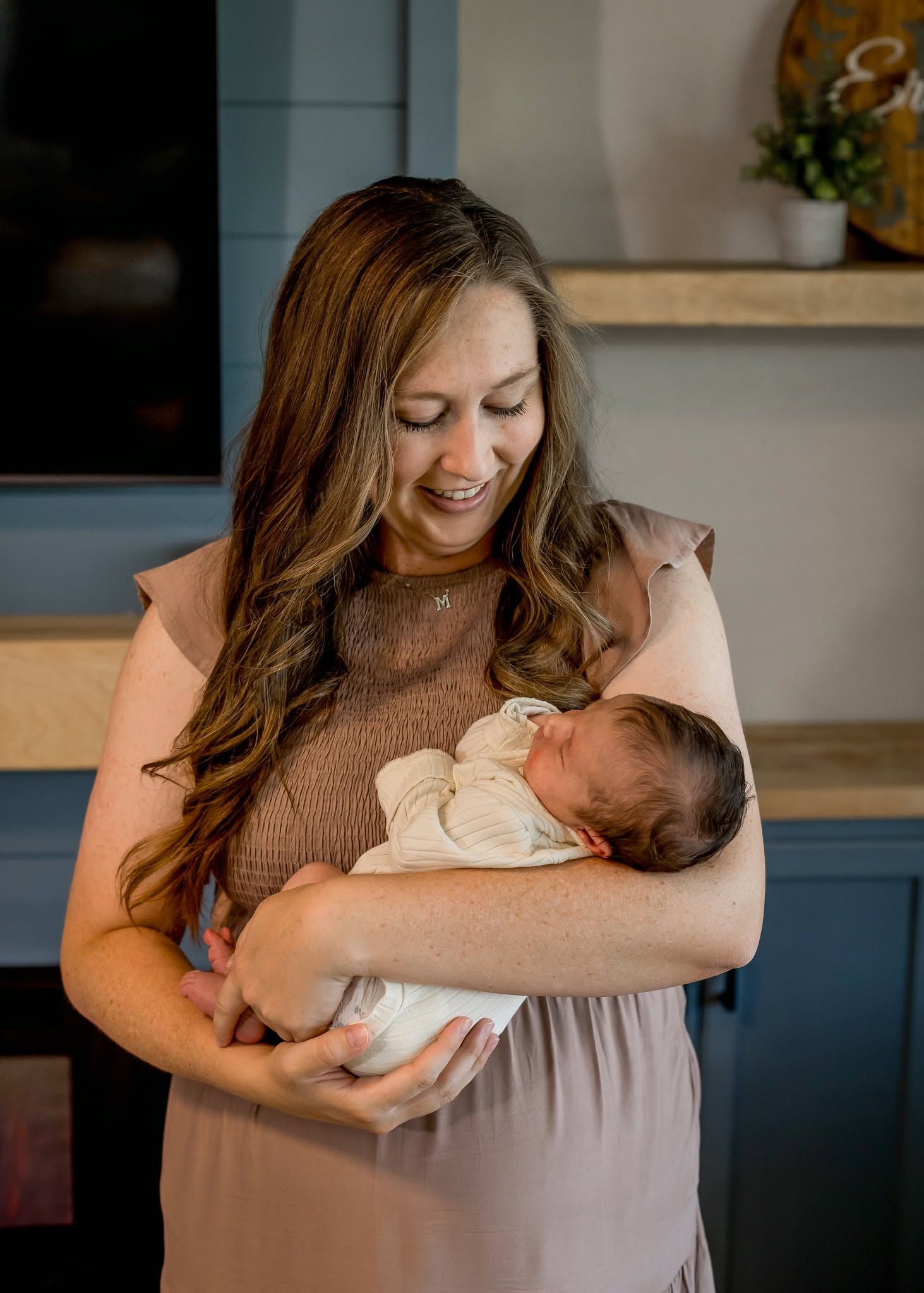 Smiling woman with long brown hair tenderly holds a sleeping newborn baby wrapped in white cloth indoors.