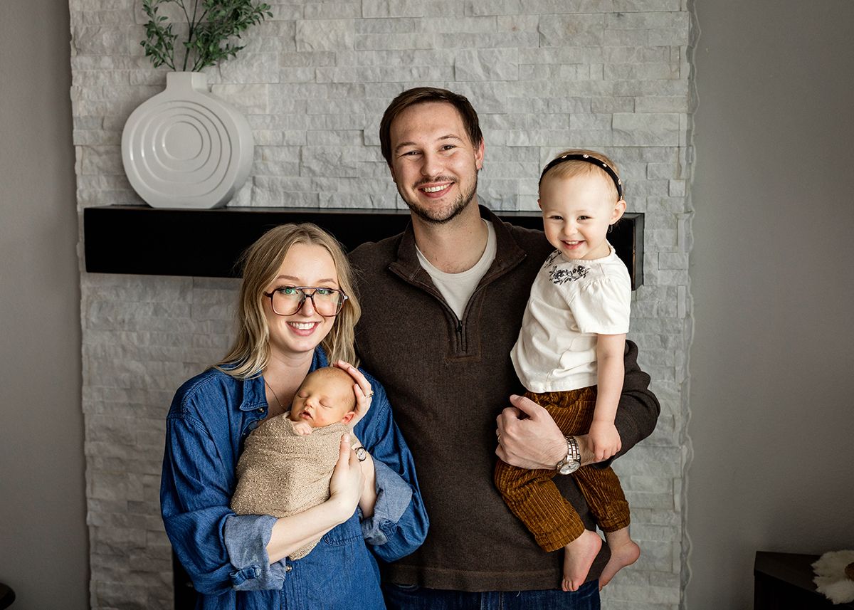 Happy family of four posing indoors, parents holding newborn and toddler in front of stone fireplace.