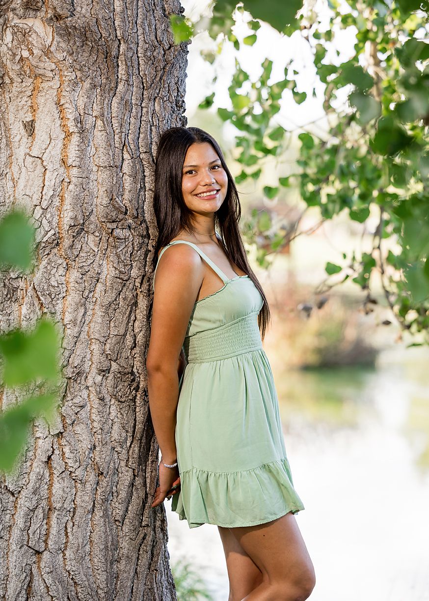 Young woman in sage green sundress smiles leaning against a tree trunk near a sunlit pond.