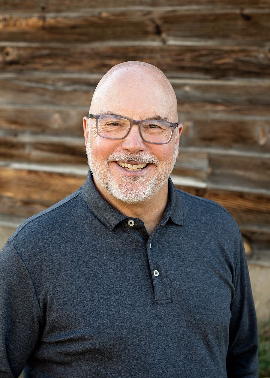 Smiling bald man with glasses and gray beard wearing a dark polo shirt, posed outdoors near a rustic wooden wall.