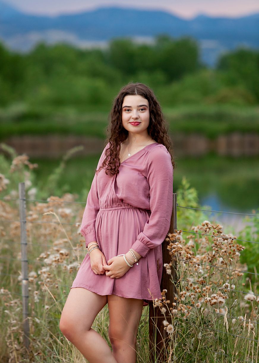 Young woman in pink dress posing outdoors near wildflowers with mountains and pond in background.