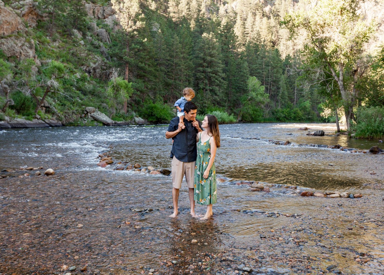 Family of three standing in a rocky river surrounded by lush green forest trees.