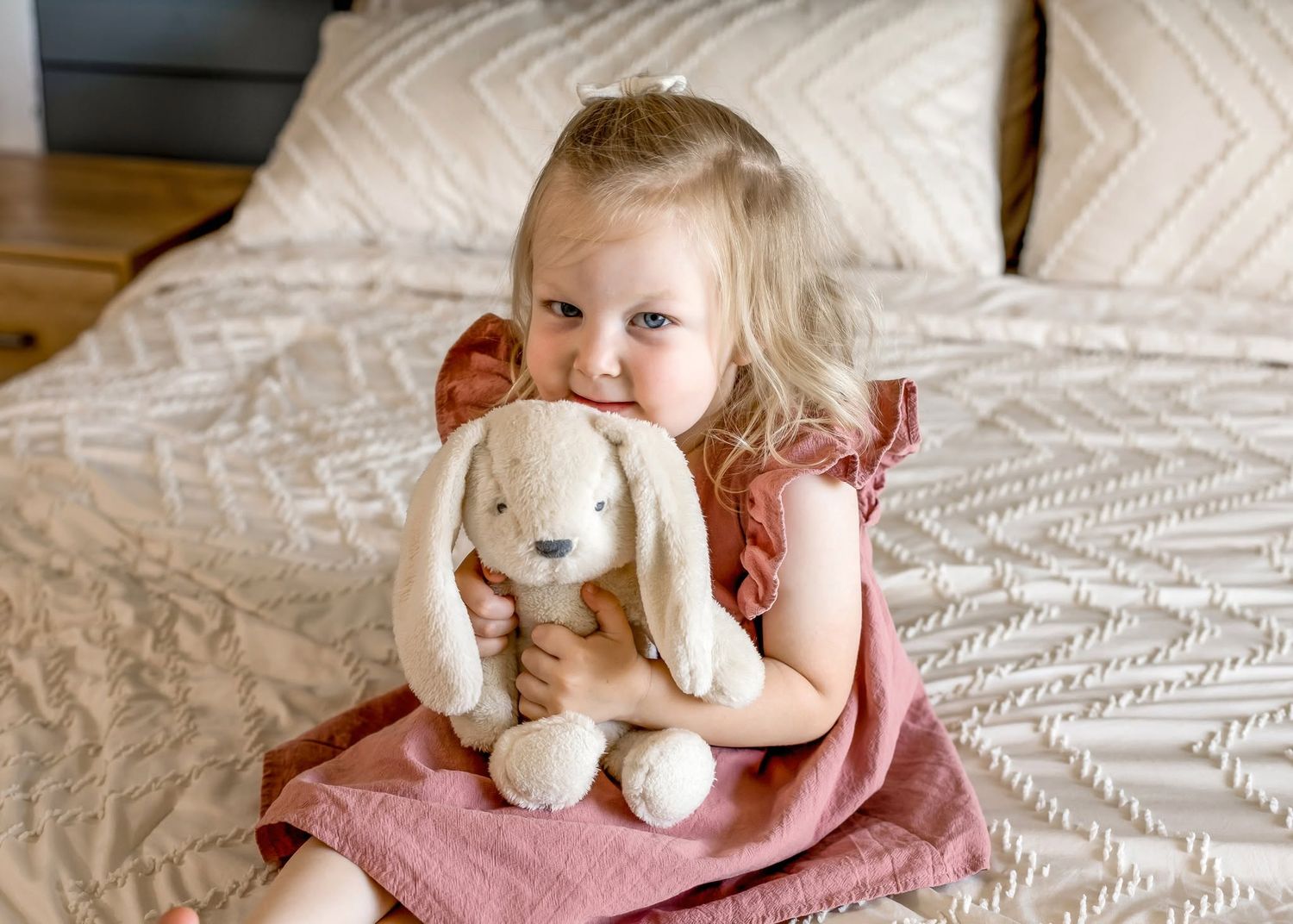 Toddler girl in pink dress hugging a cream plush bunny stuffed animal while sitting near a bed.