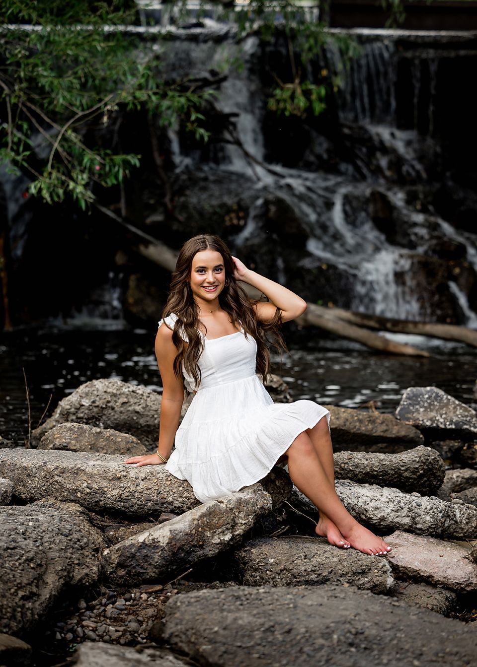 Smiling young woman in white dress sits barefoot on rocks in front of a scenic waterfall.