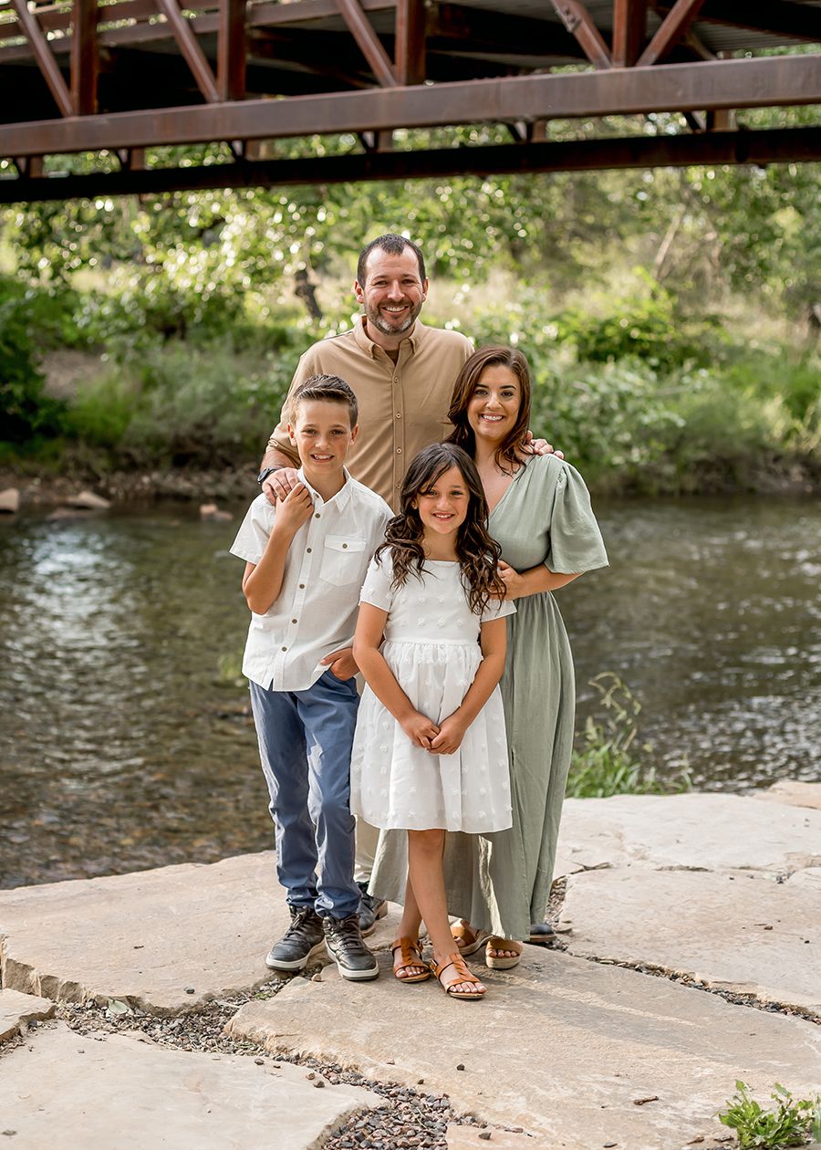 Family of four posing by a creek under a bridge, parents with two children in coordinating outfits.