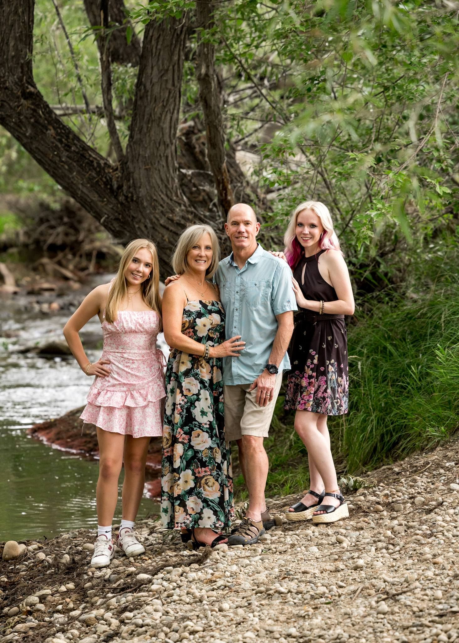 Family of four smiling together on a rocky creek bank surrounded by trees and green foliage.