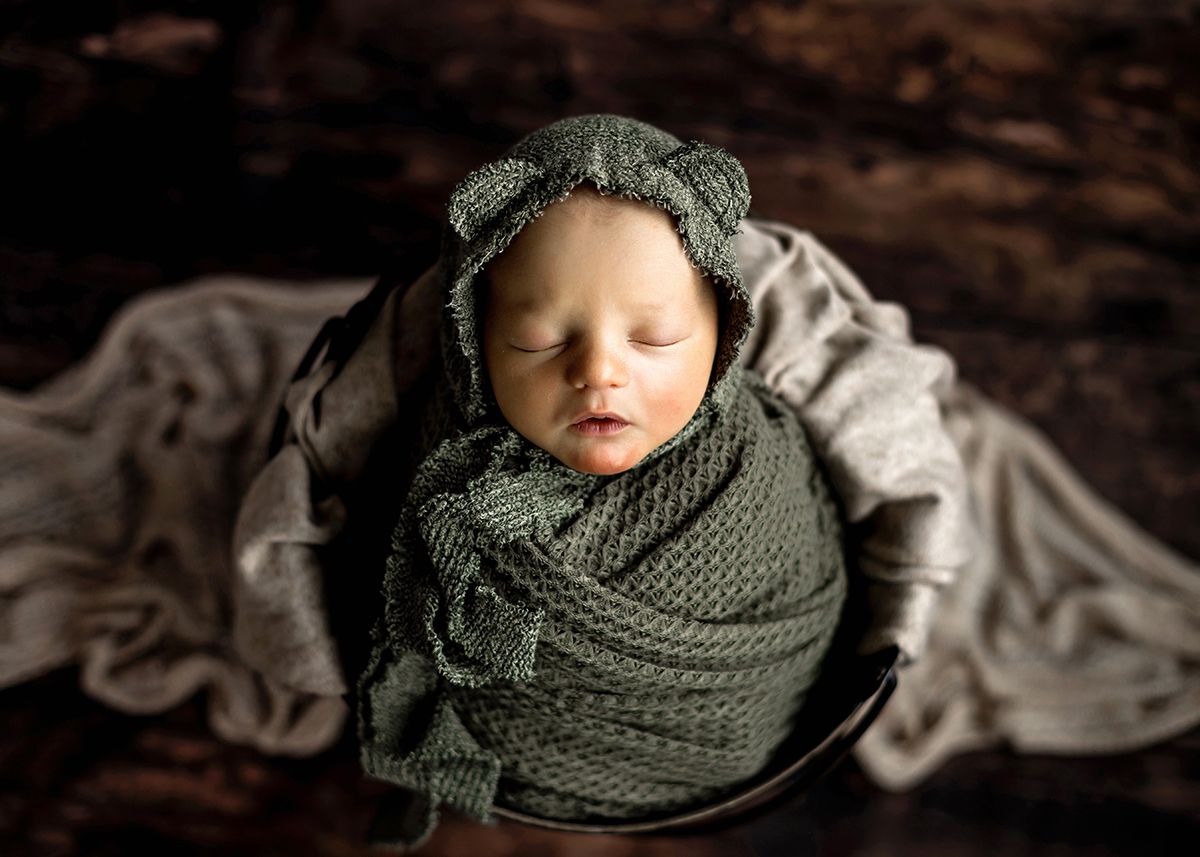 Newborn baby swaddled in green knit wrap wearing bear bonnet sleeping in wooden bowl.