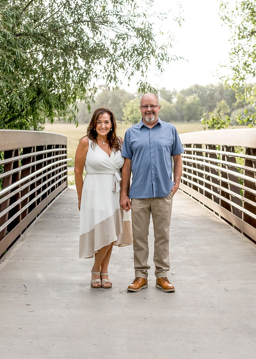 Couple smiling and holding hands on a wooden bridge surrounded by lush greenery on a sunny day.