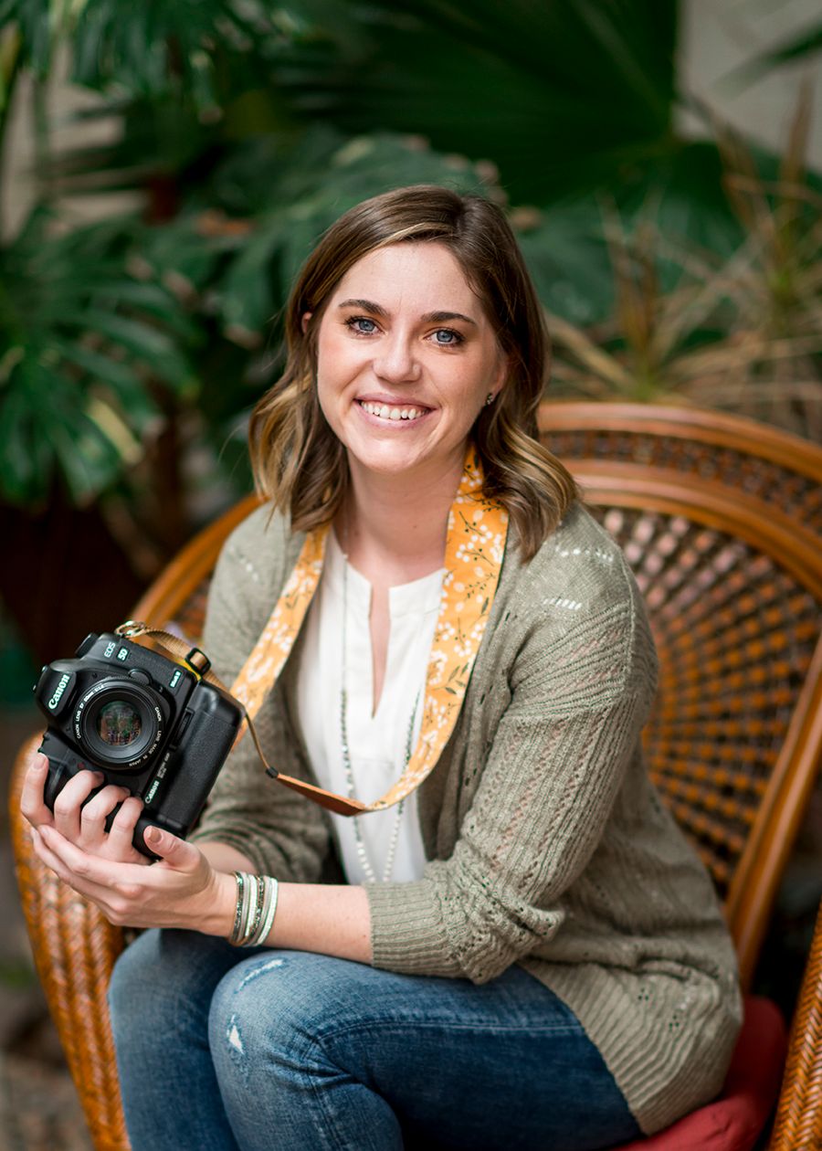 Smiling woman holding a camera while seated in a wicker chair among tropical plants.