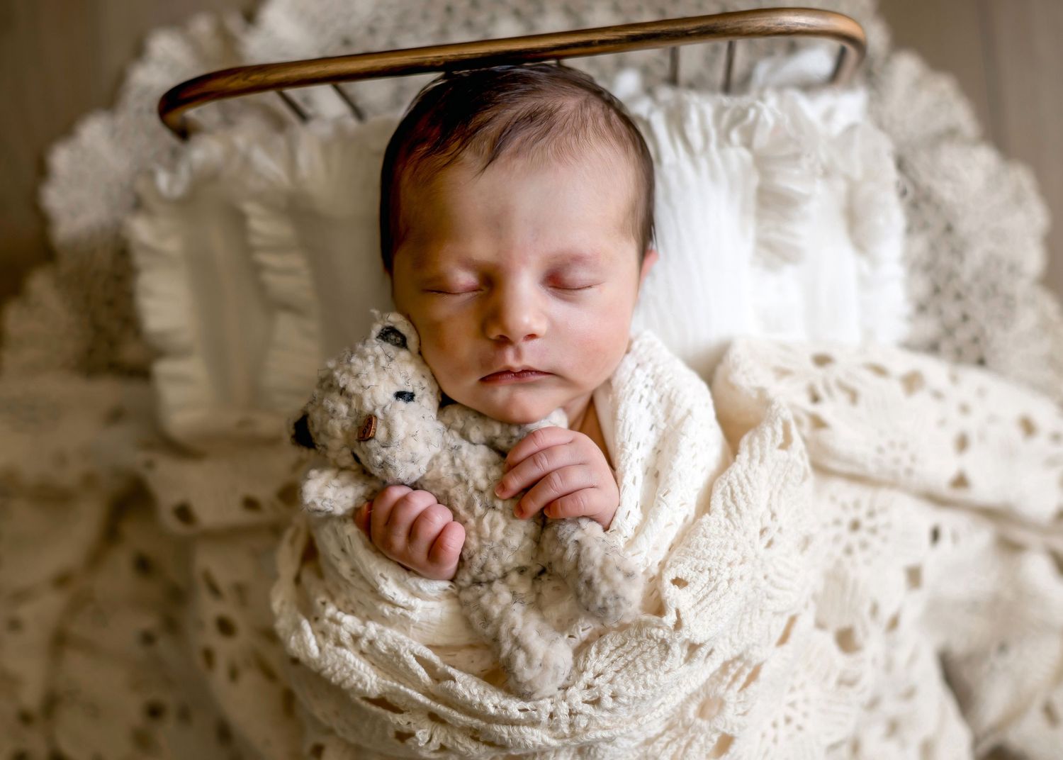 Sleeping newborn baby wrapped in cream lace swaddle cuddling a small teddy bear.