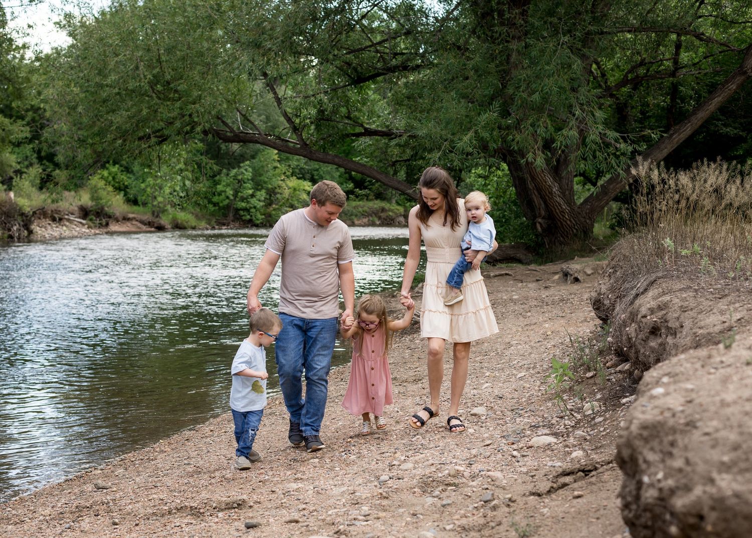 Family of five strolling along a peaceful riverbank path lined with green trees.