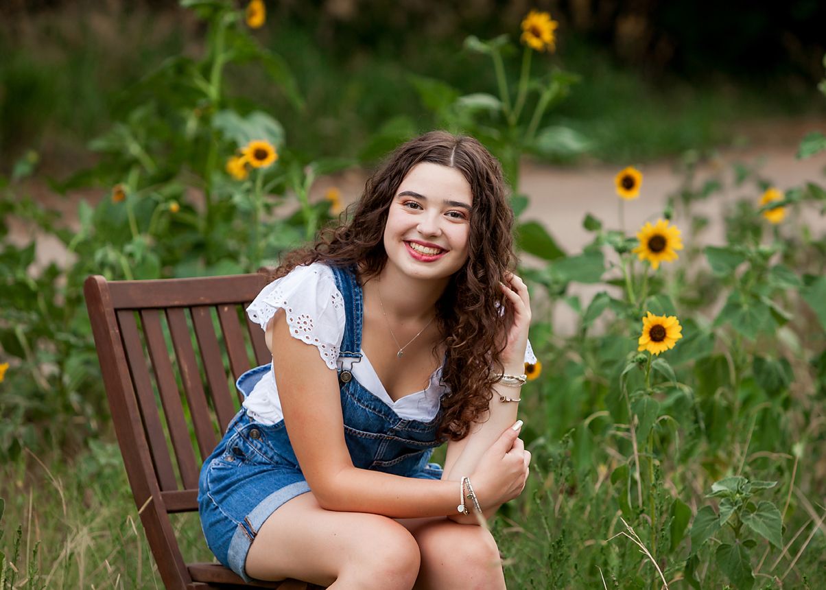 Smiling teen girl in denim overalls sits on wooden chair surrounded by sunflowers for senior portrait.
