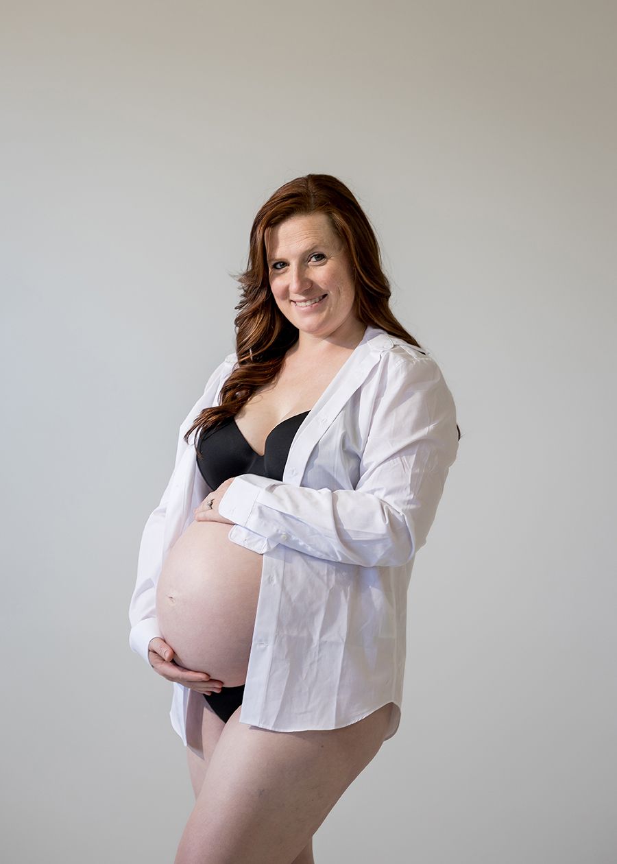 Smiling pregnant woman in open white shirt and black bra cradling her baby bump in a studio portrait.