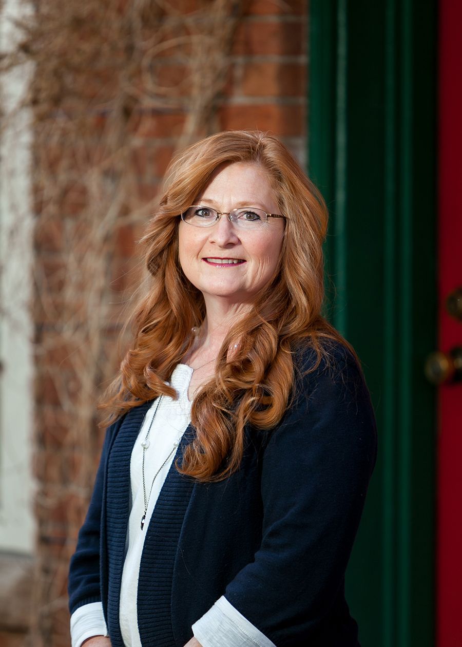 A smiling woman with red wavy hair and glasses wearing a navy cardigan stands outdoors near a brick building.