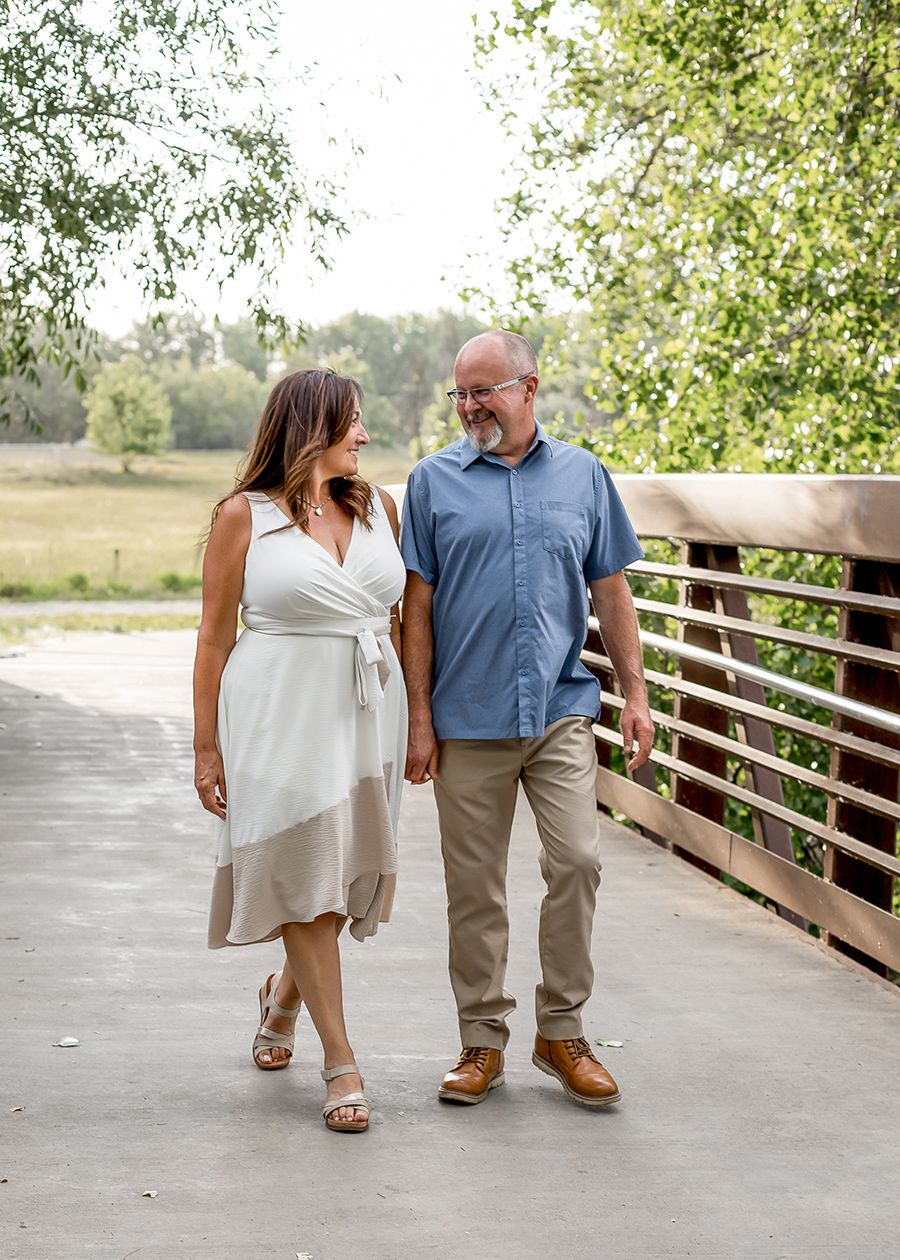 Couple walking hand in hand on a bridge, woman in white dress, man in blue shirt, surrounded by greenery.