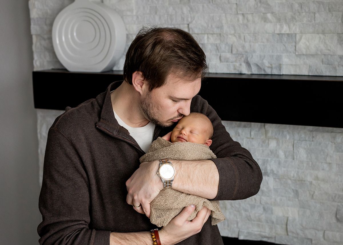 A father tenderly kisses his swaddled newborn baby while standing near a stone fireplace.