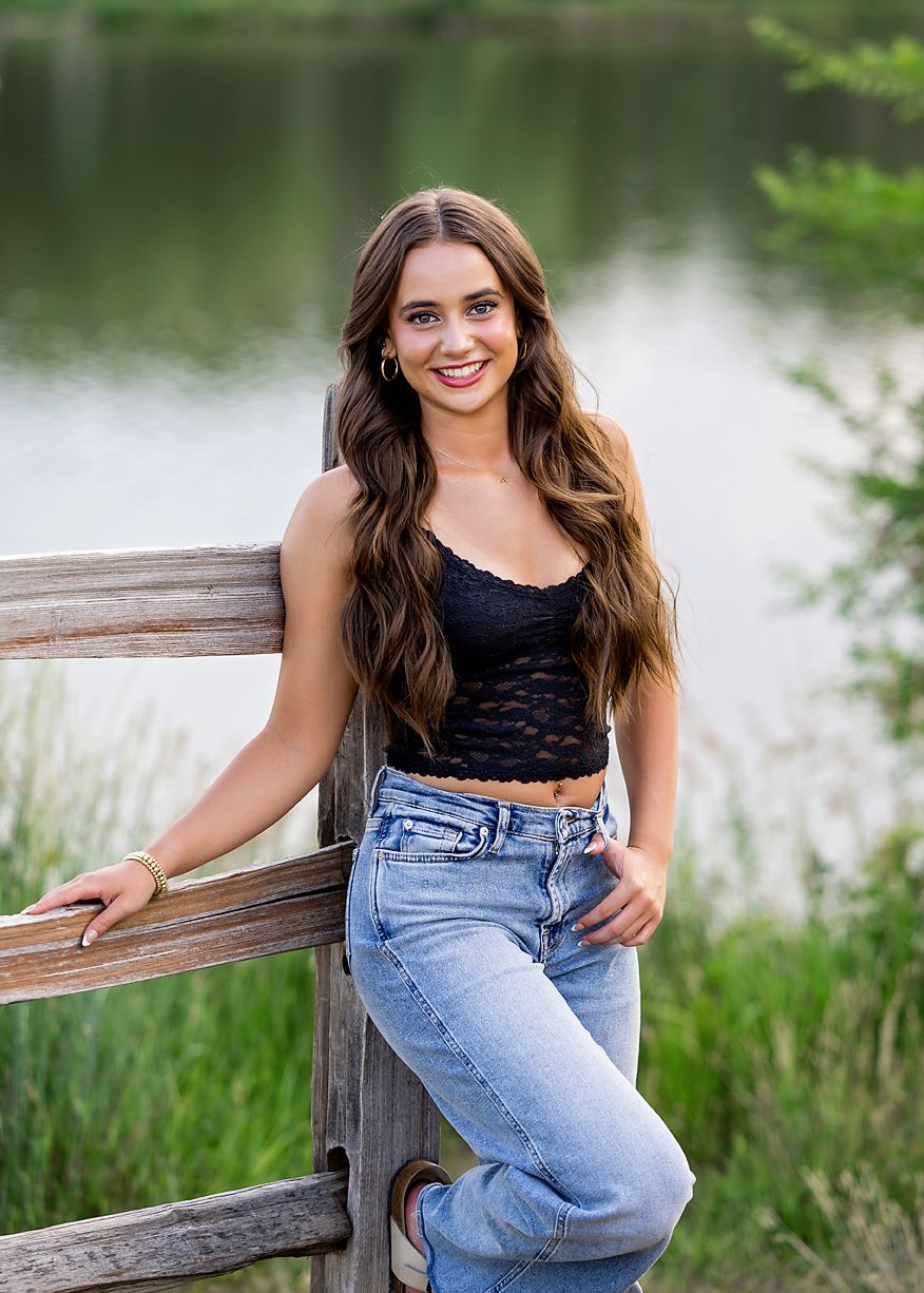 Young woman in black crop top and jeans smiling by a wooden fence near a pond.