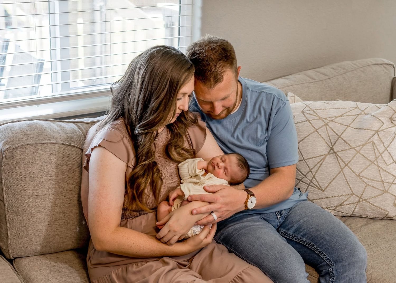 A couple sits on a couch gazing lovingly at their newborn baby, sharing a tender family moment.