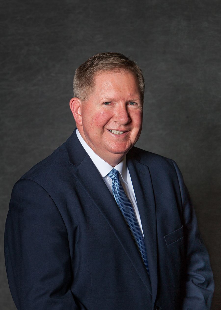 Professional headshot of a smiling middle-aged man in a navy suit and light blue tie against a gray background.