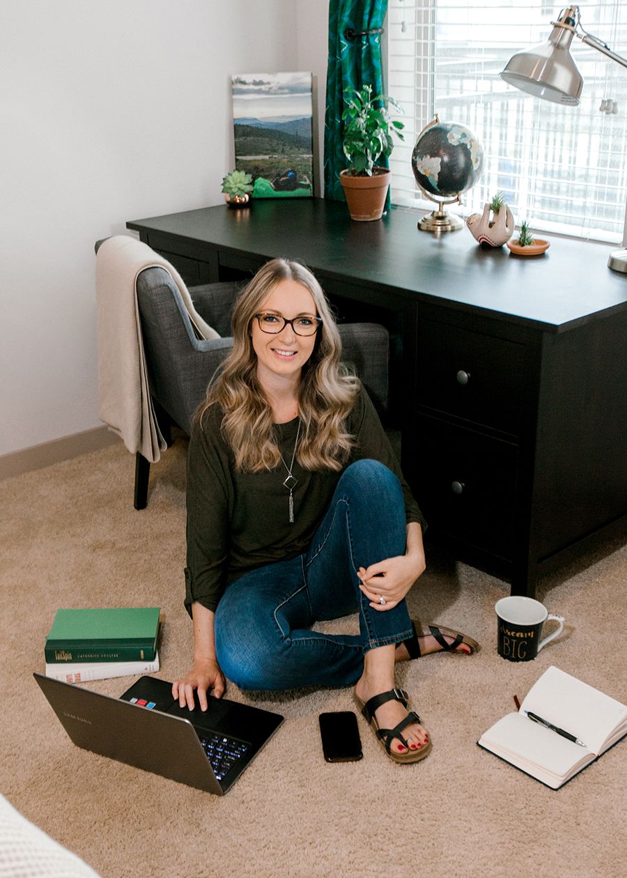 Woman with glasses sitting on floor by dark desk with laptop, phone, notebook, and coffee mug.