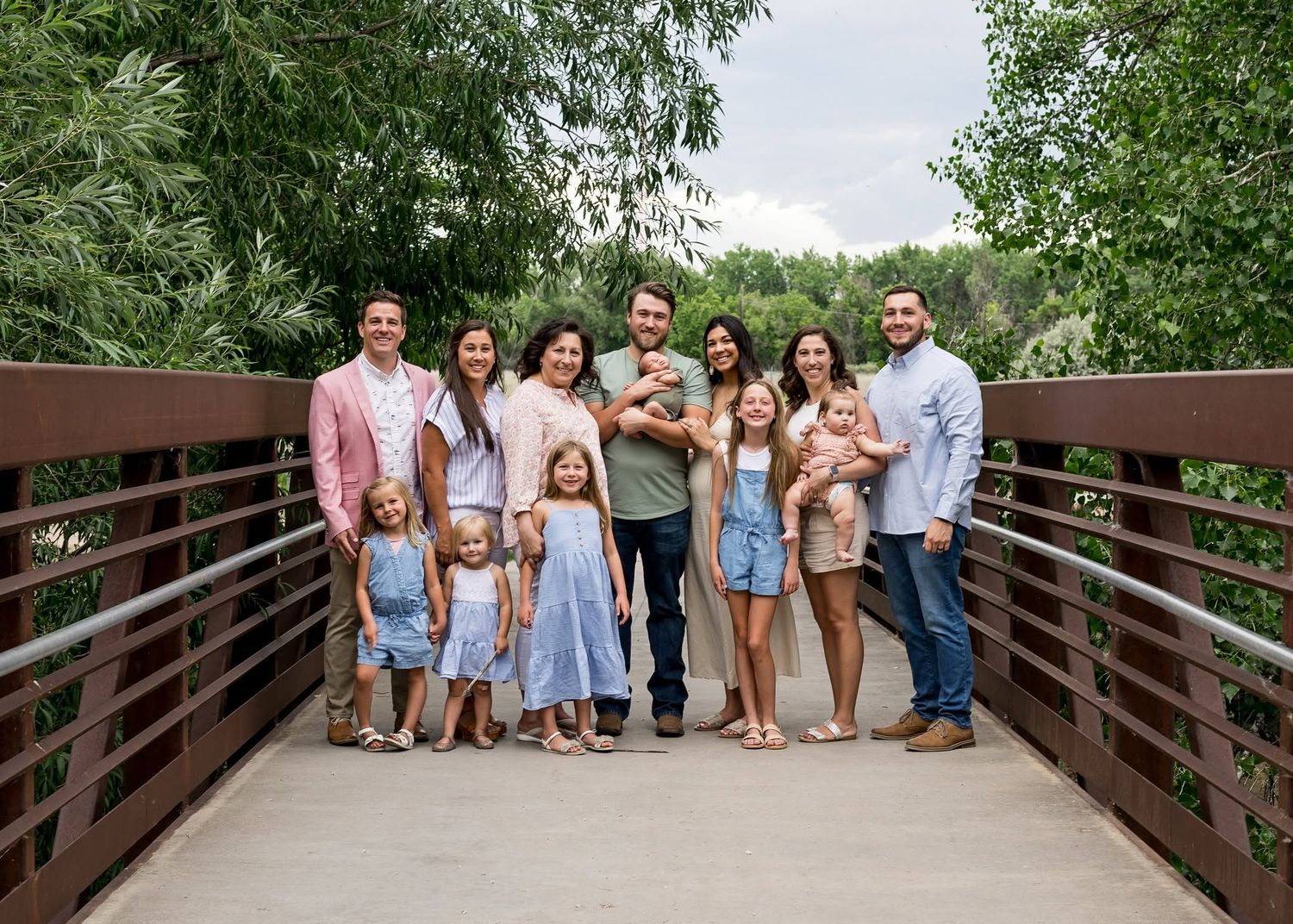 Large family posing together on a wooden bridge surrounded by lush green trees on a sunny day.