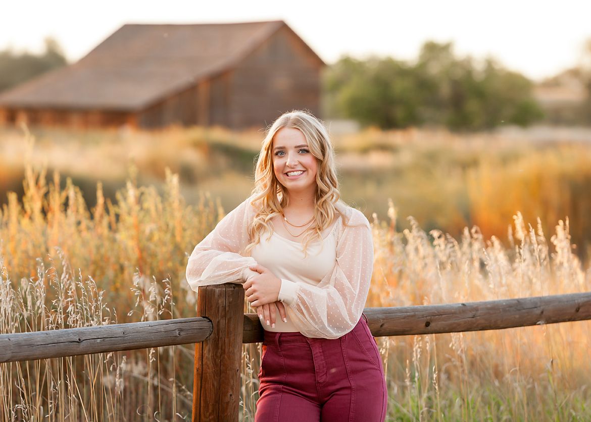 Smiling blonde teenage girl in pink top and burgundy pants leaning on rustic fence in golden field.