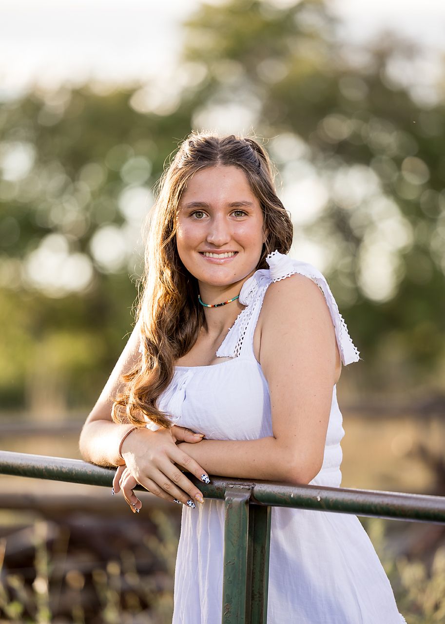 Young woman in white dress leaning on a green fence, smiling outdoors with golden hour sunlight.