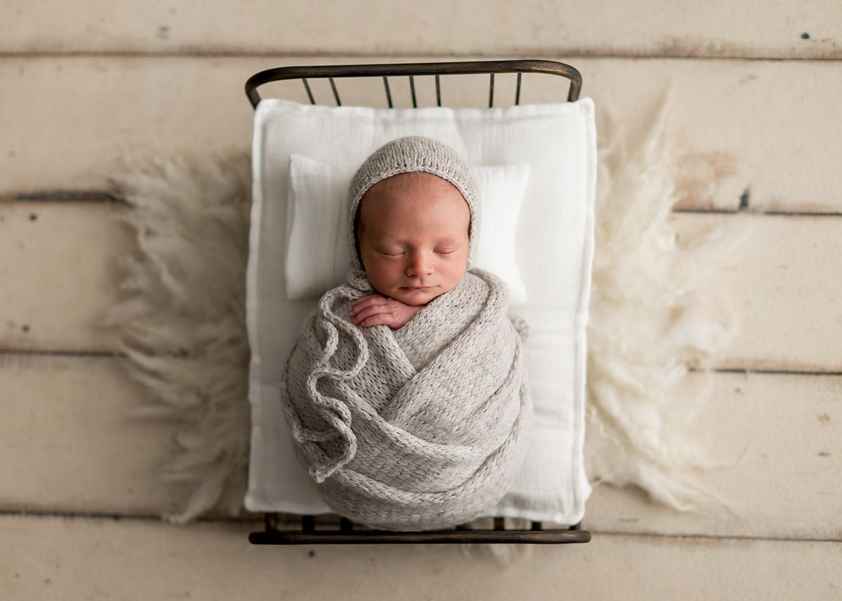 Newborn baby swaddled in a gray knit wrap and bonnet, sleeping peacefully in a tiny vintage metal bed.