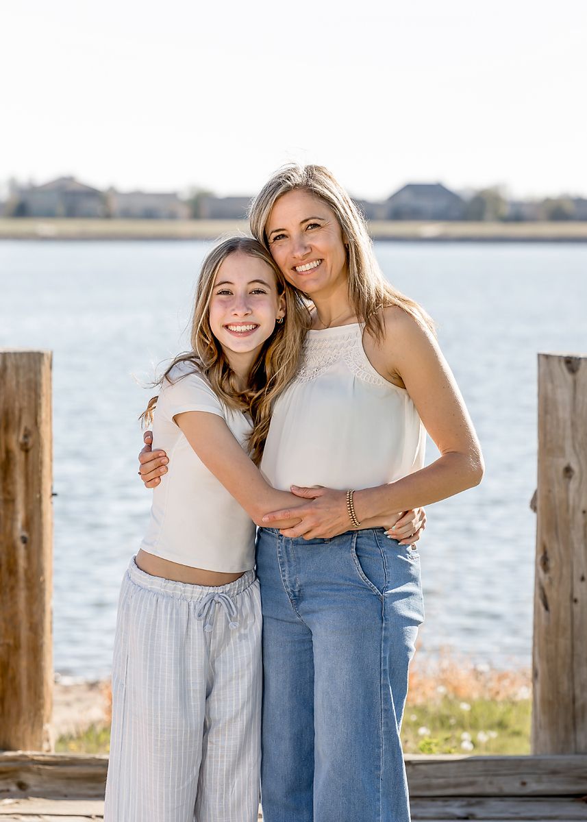 A smiling mother and daughter embrace on a sunny lakeside wooden dock.