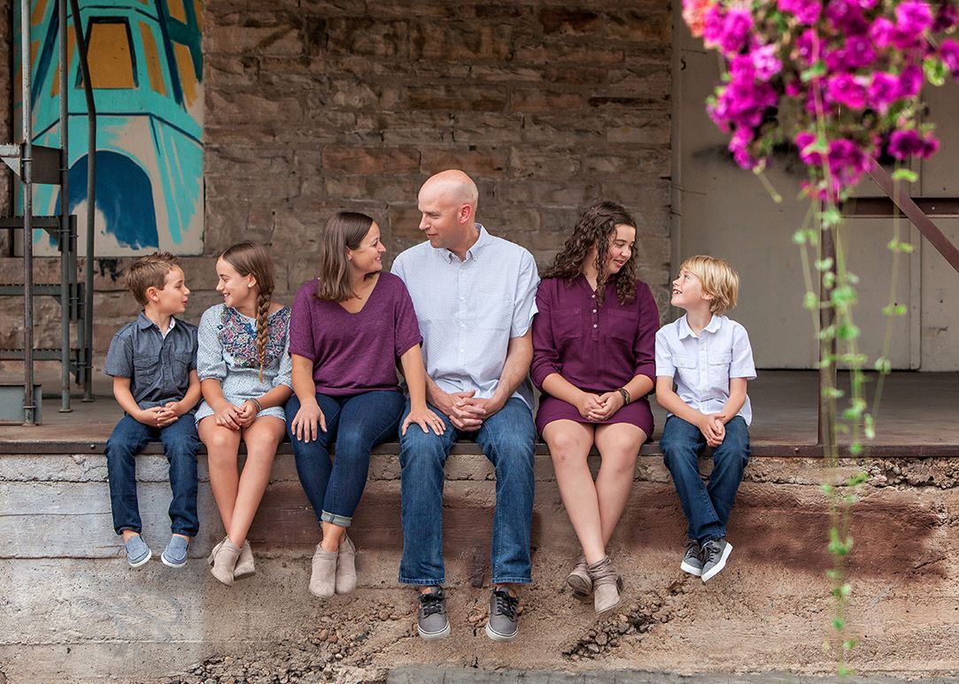 Family of six posing on rustic stone steps beside vibrant pink bougainvillea in coordinating purple outfits.