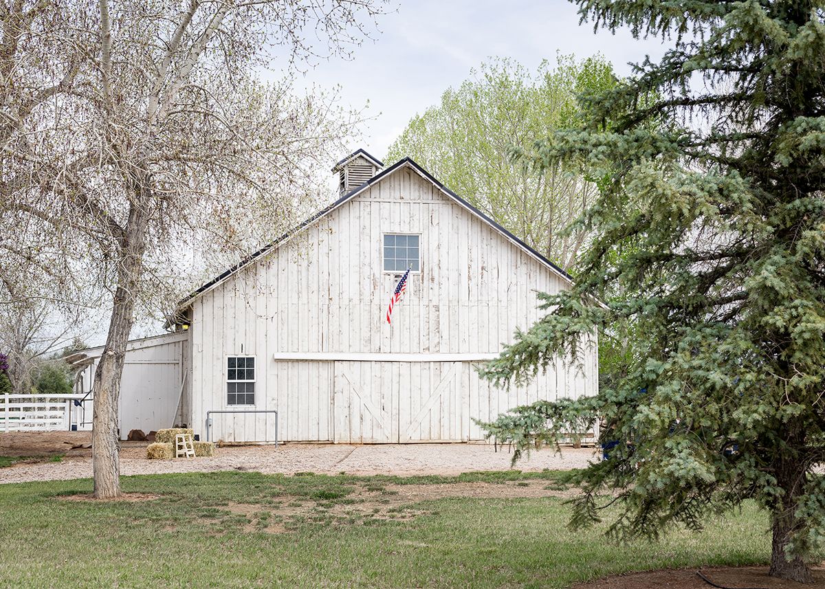 Rustic white barn with an American flag displayed on the side surrounded by green trees.