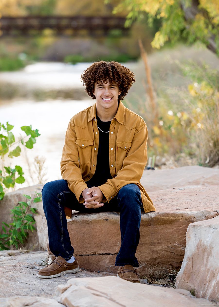 Young man with curly hair sits smiling on a rock outdoors, wearing a mustard jacket and jeans near a river.