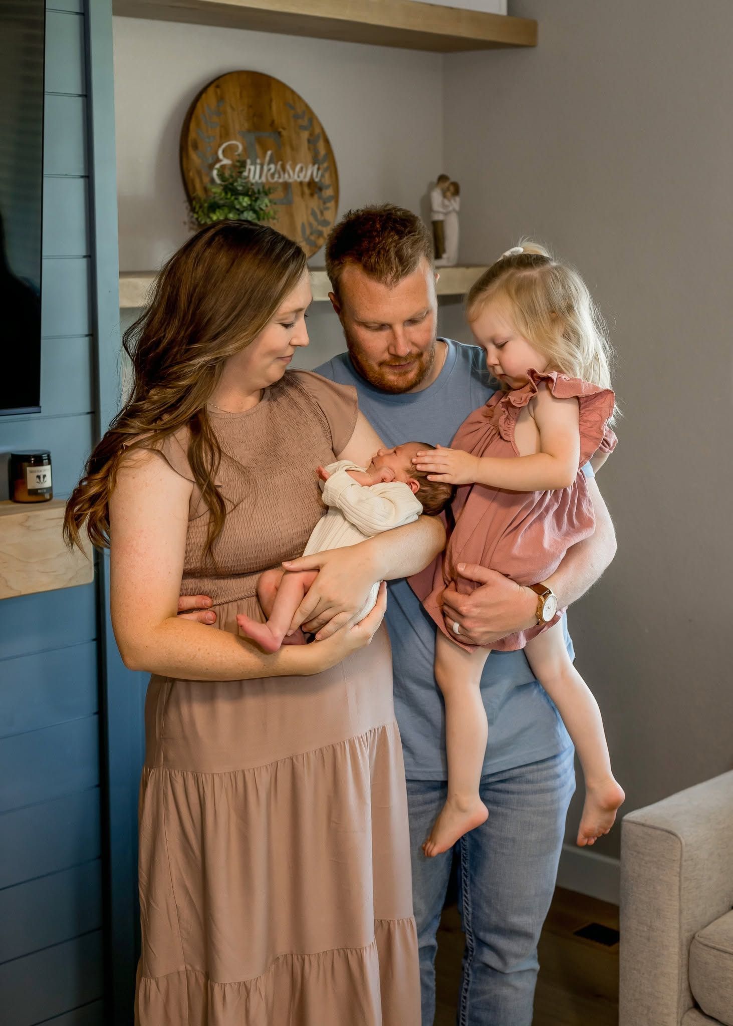 A young family of four admires their newborn baby, with parents and toddler daughter gathered together in a cozy home setting.