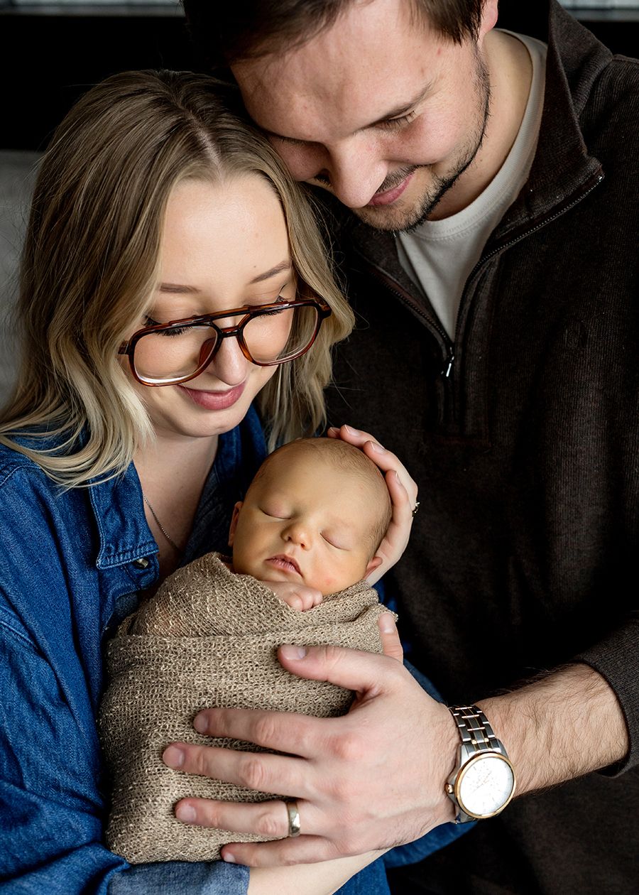 A couple tenderly gazes at their sleeping newborn baby wrapped in a burlap swaddle during a newborn portrait session.