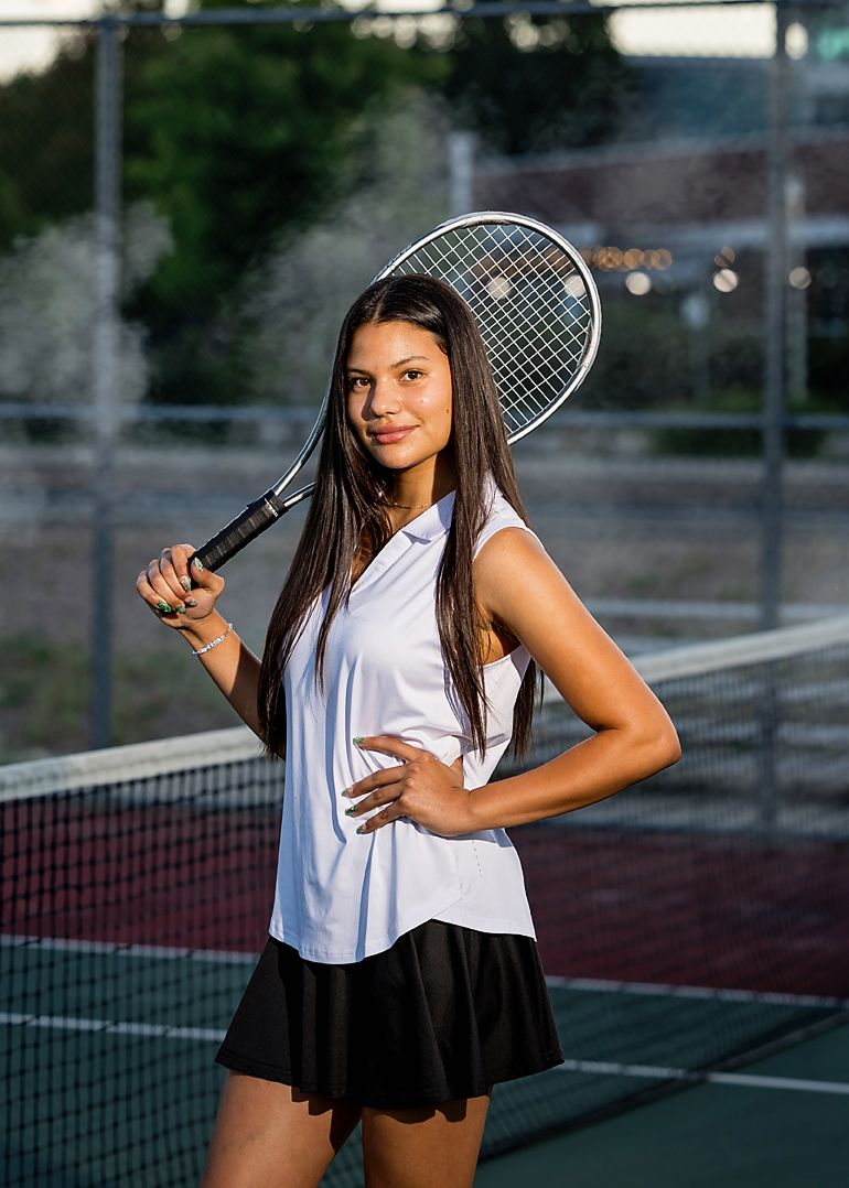Young female tennis player poses confidently on court holding racket, wearing white top and black skirt.
