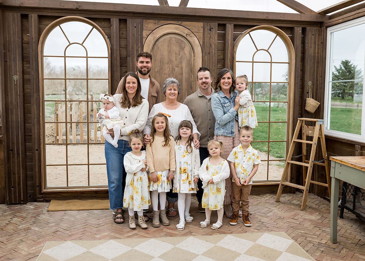 Large multigenerational family portrait with grandparents, parents, and children posing in a rustic wooden greenhouse.