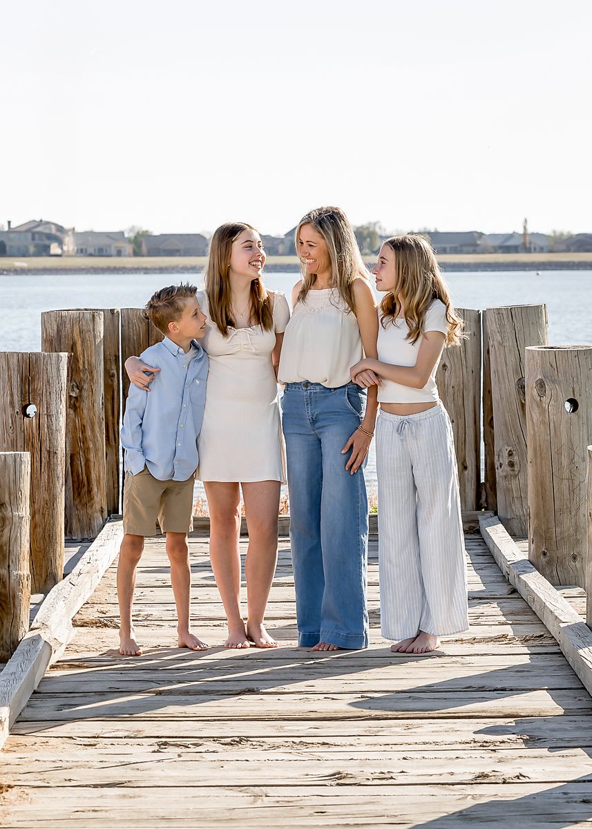 Four siblings laugh together on a rustic wooden pier by the water.