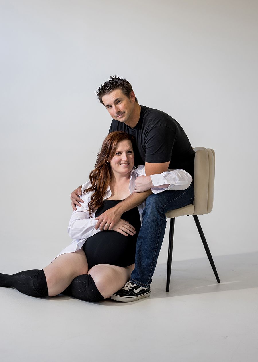 A couple poses for a maternity photo, woman sitting on floor in black outfit, man in black shirt seated on chair behind her.