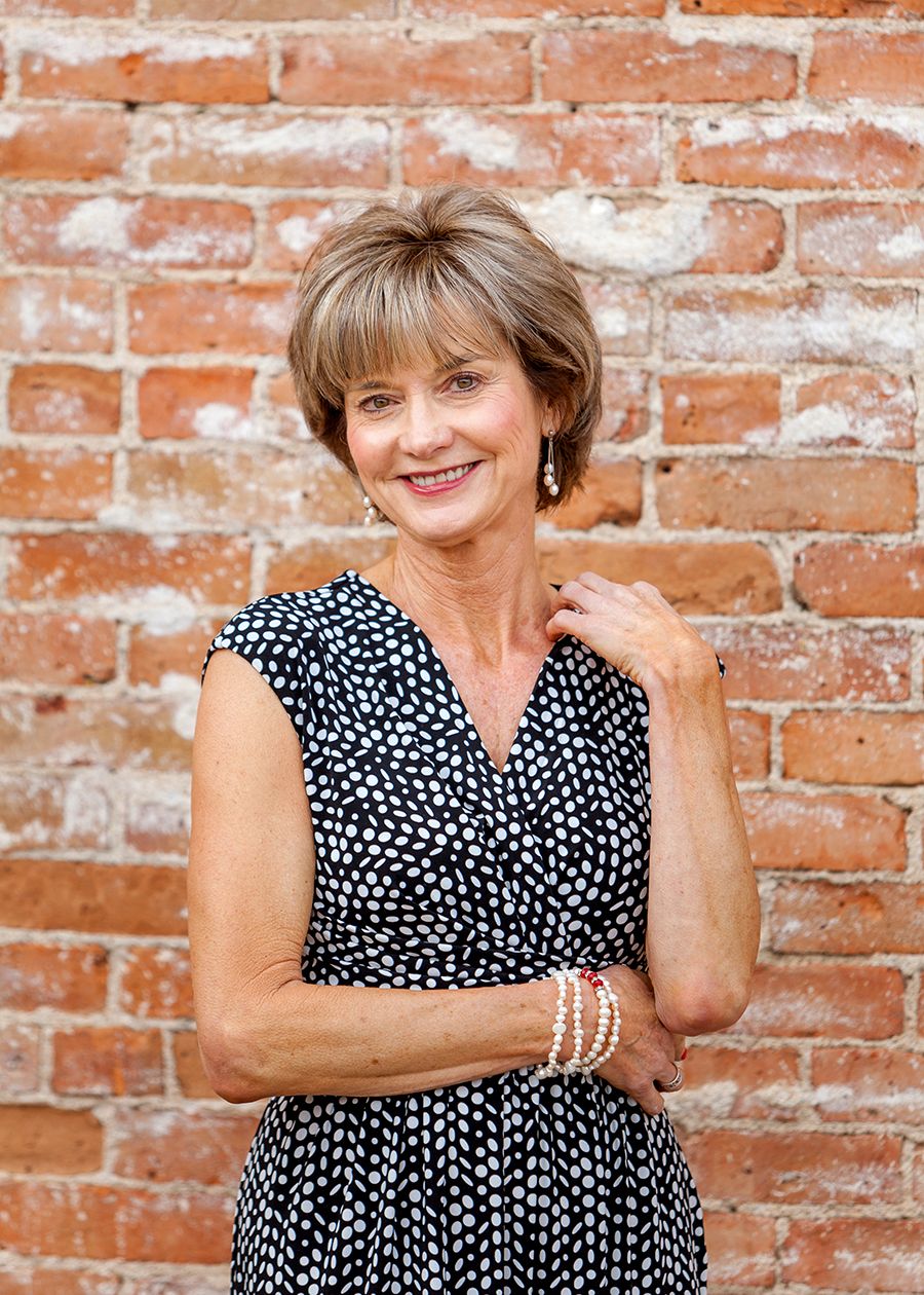 Smiling middle-aged woman in polka dot dress posing against rustic red brick wall wearing pearl bracelet and earrings.
