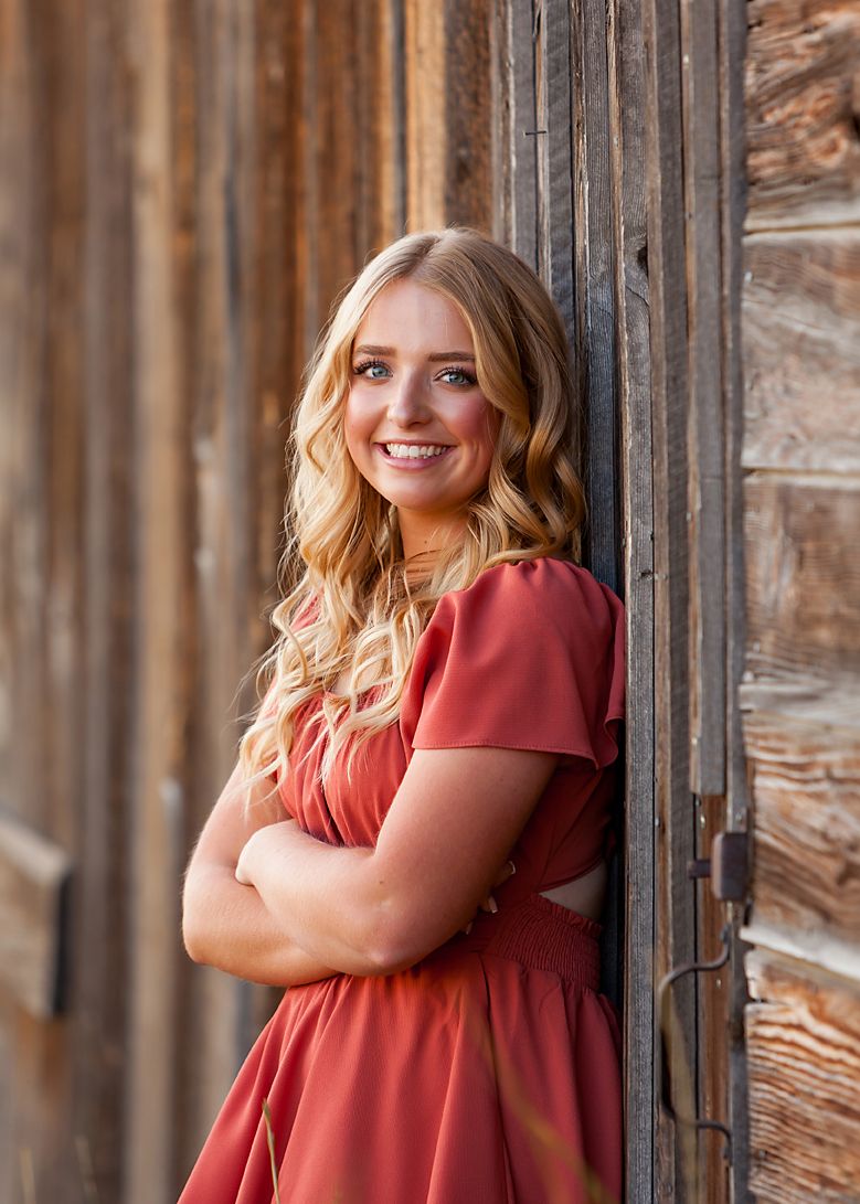 Young woman in red dress with crossed arms smiling against rustic wooden wall in senior portrait photo.