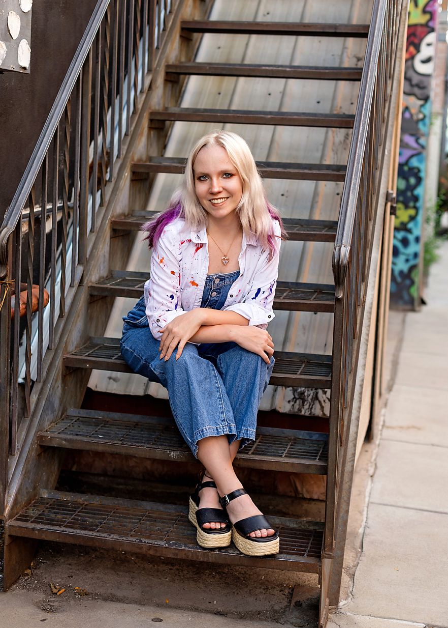 Smiling blonde teen girl with pink highlights sits on metal stairs near colorful graffiti wall.